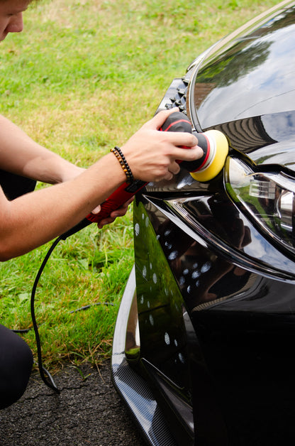 Person polishing a black car with a polisher on a grassy background